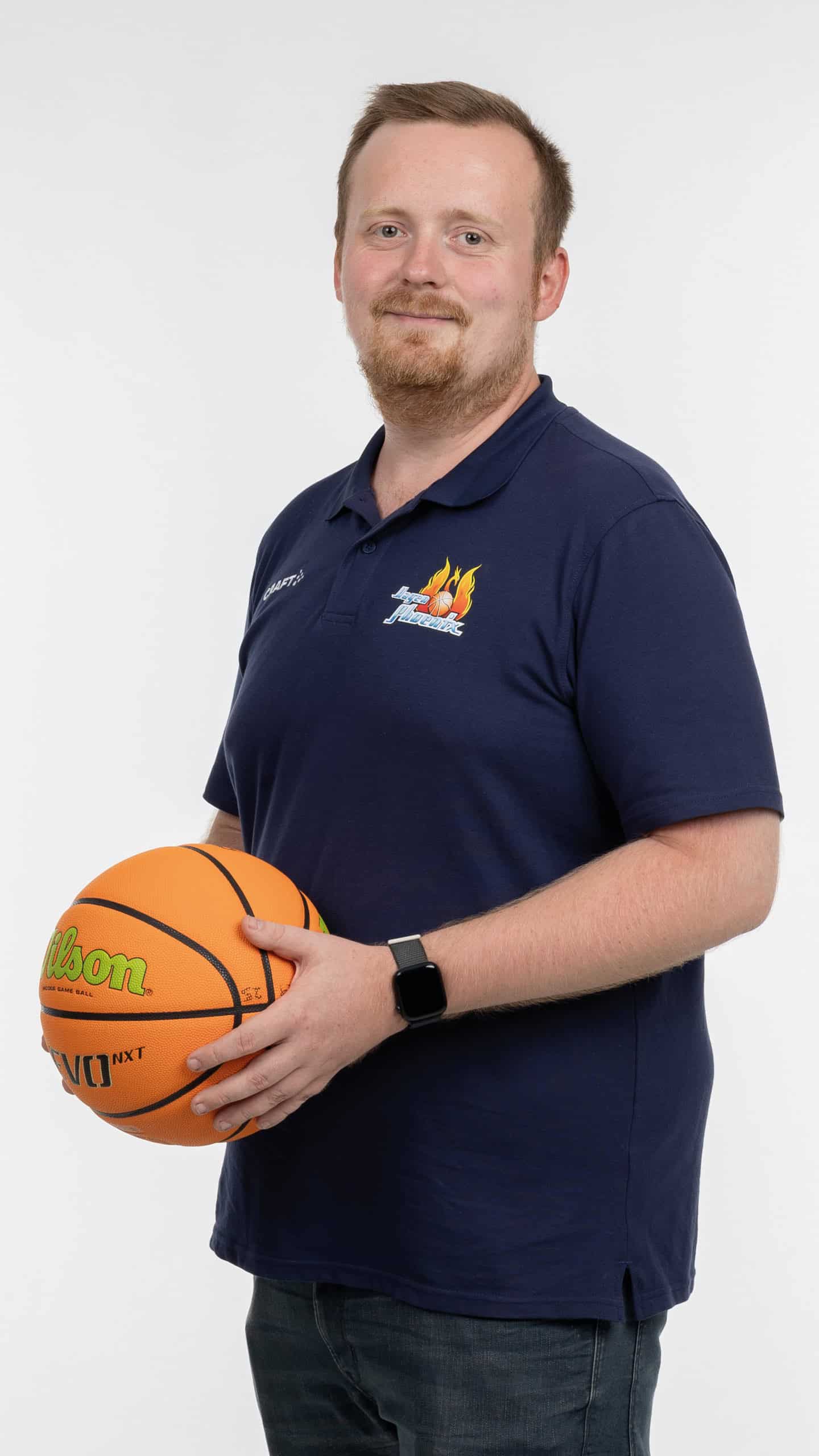 Mann in blauem Polohemd, lächelnd, hält einen Basketball, weißer Hintergrund.