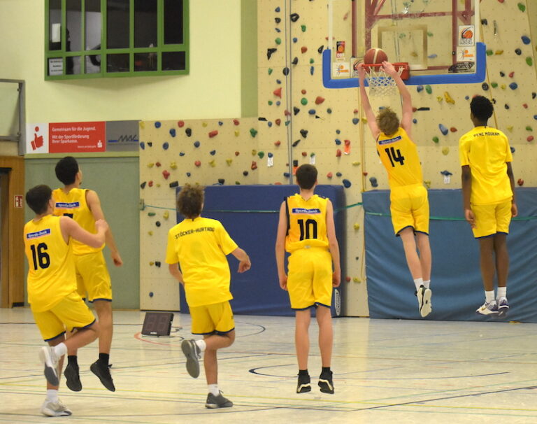 Sechs Basketballspieler im gelben Trikot auf dem Spielfeld, einer macht einen Dunk, Turnhalle.