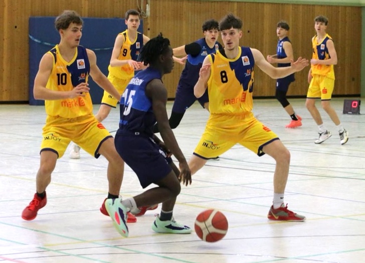Fünf Basketballspieler im Spiel. Zwei im gelben Trikot verteidigen, einer im blauen dribbelt.