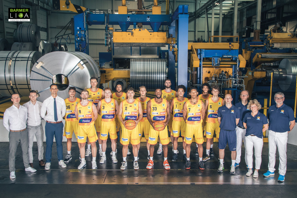 Gruppe von Basketballspielern in gelben Trikots in einer Fabrikhalle, alle lächeln in die Kamera.