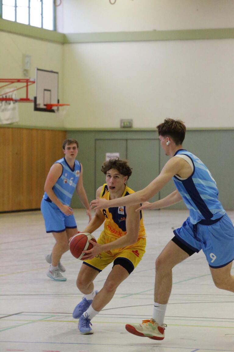Ein junger Mann im gelben Trikot dribbelt einen Basketball, verfolgt von Gegenspielern in Blau.