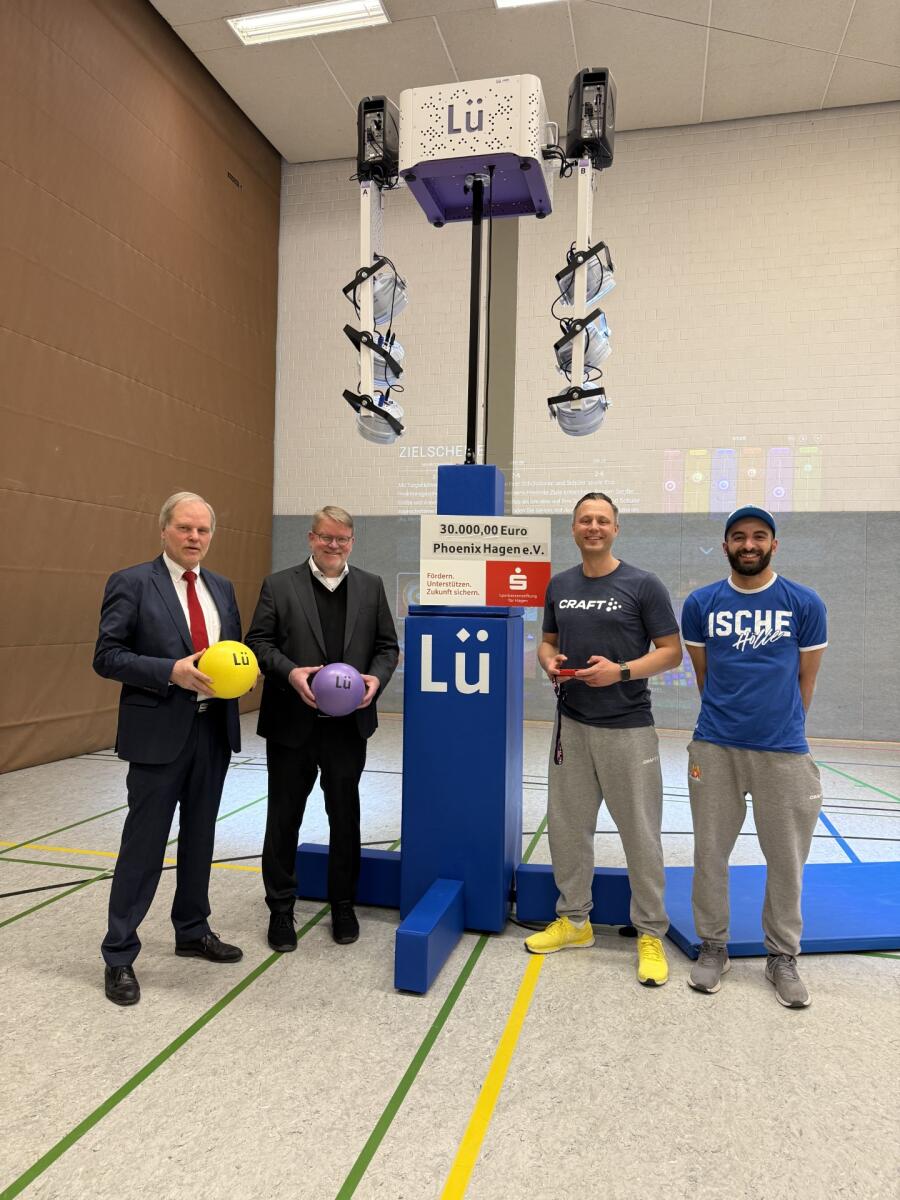 Vier Männer posieren lächelnd in einer Sporthalle mit einem Scheck und bunten Trainingsutensilien.