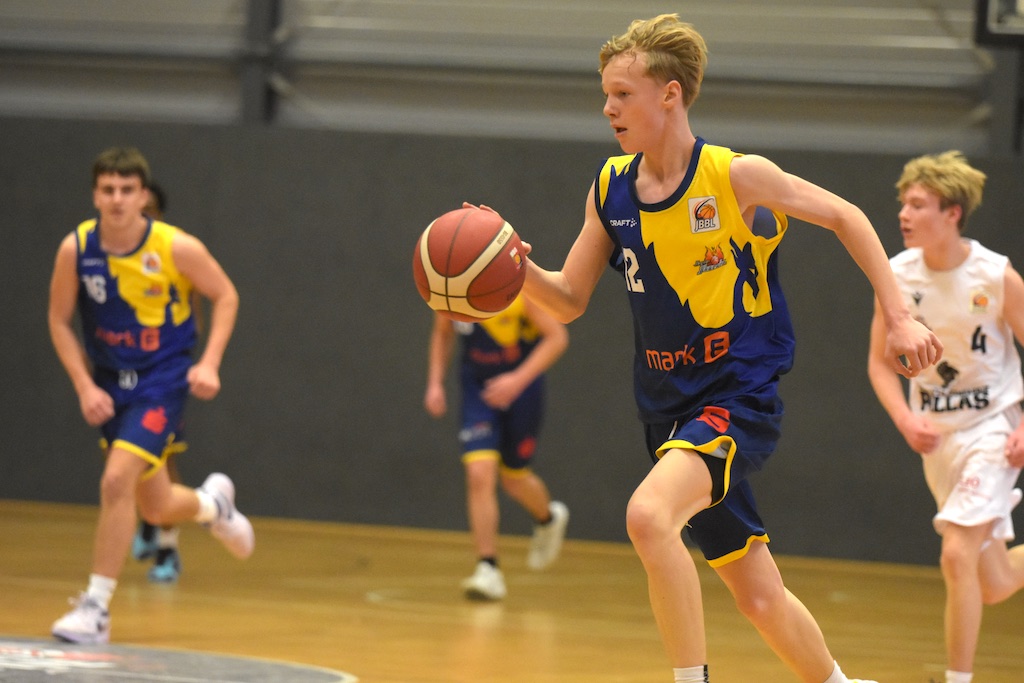 Ein junger Mann spielt Basketball, fokussiert, in blau-gelbem Trikot auf einem Hallenplatz.