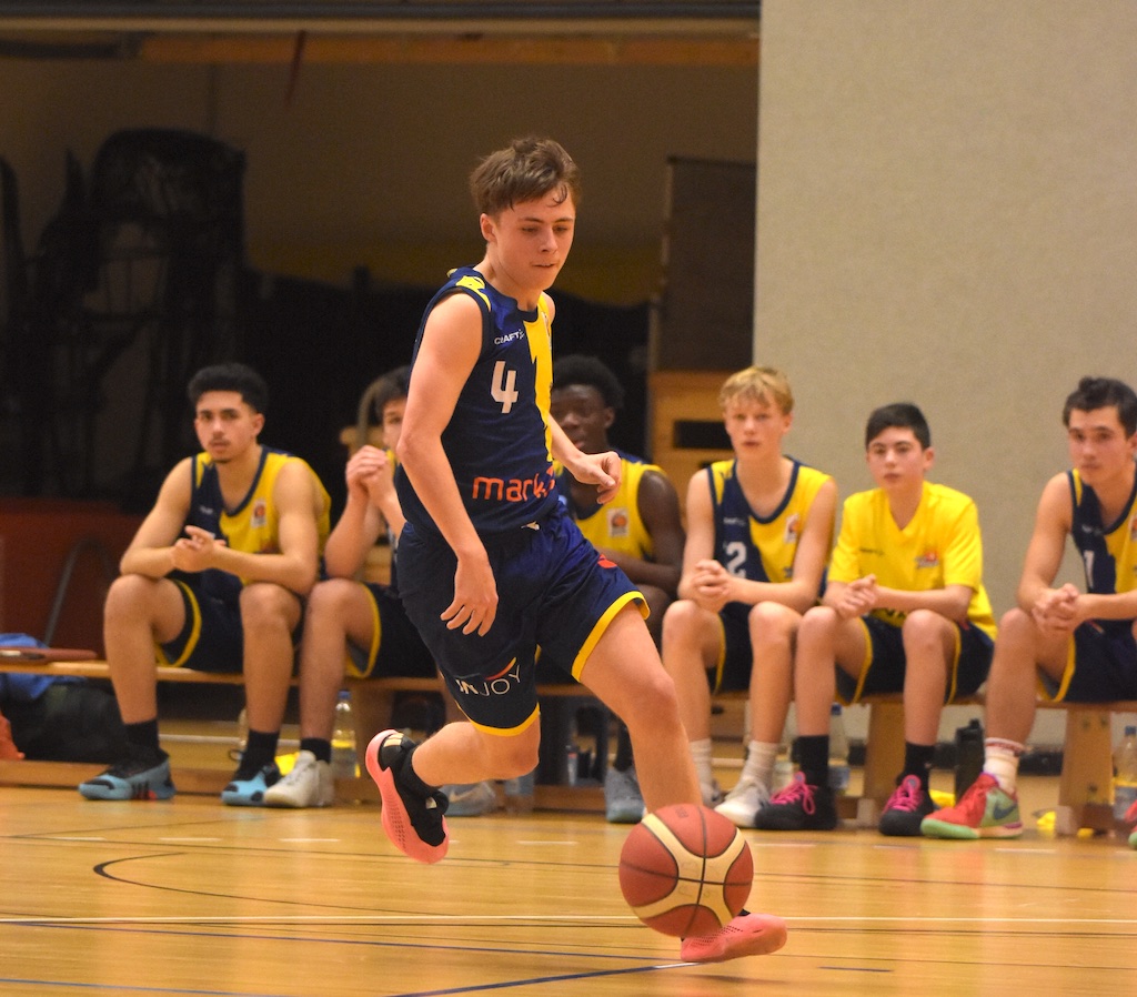 Ein Jugendlicher spielt Basketball; blaue Uniform, konzentrierten Ausdruck, Zuschauer auf der Bank.