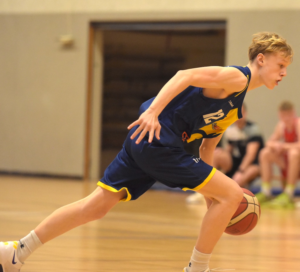 Junger Mann spielt Basketball, konzentrierter Ausdruck, blau-gelbes Trikot, hallenartiger Raum.