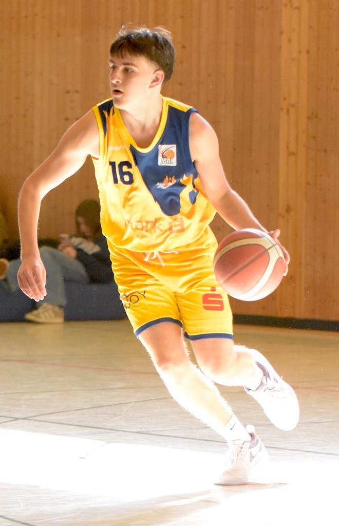Ein Basketballspieler im gelben Trikot dribbelt konzentriert einen Ball in einer Halle.