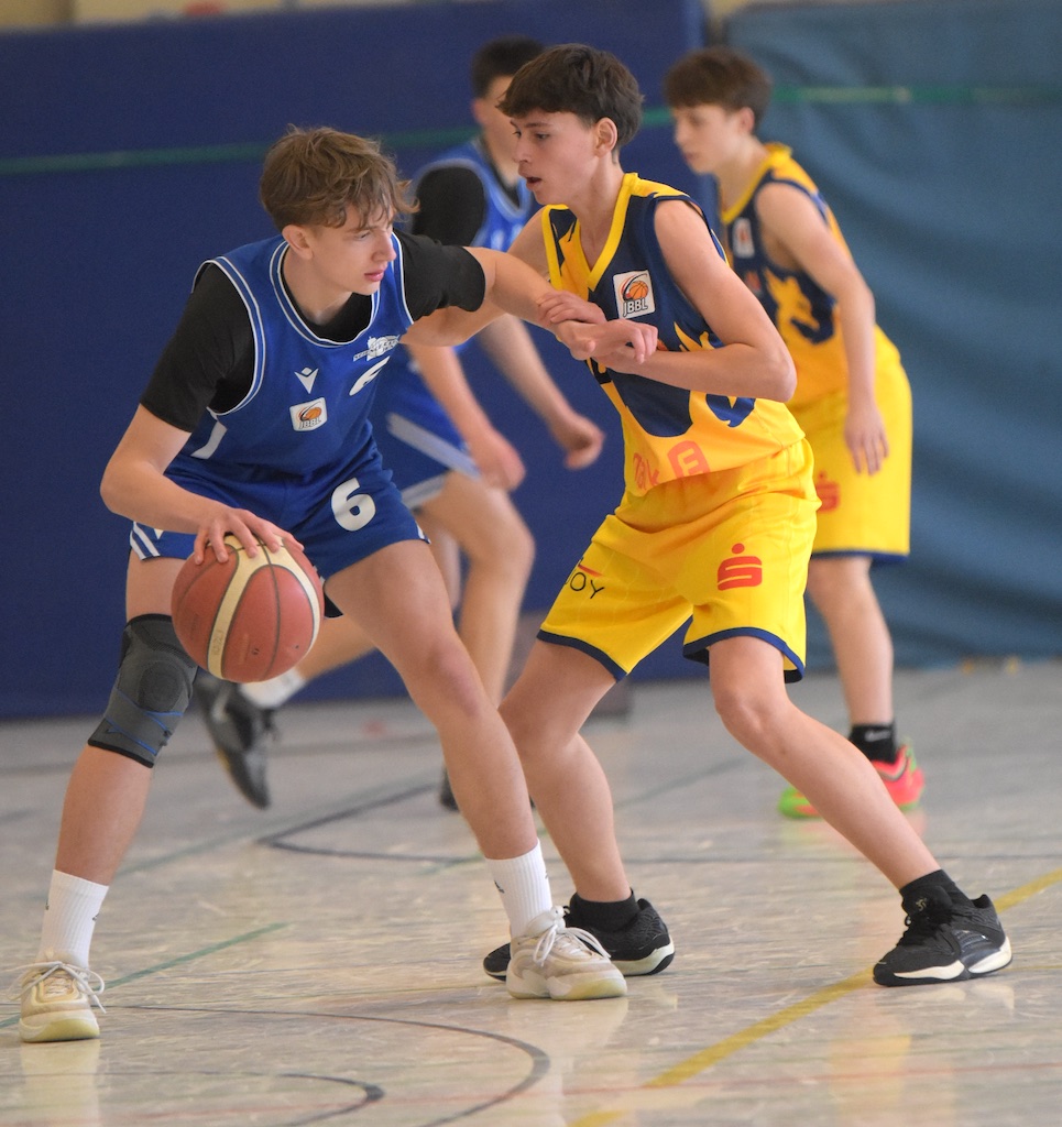 Ein Basketballspieler im blauen Trikot dribbelt, verteidigt von einem Spieler im gelben Trikot.