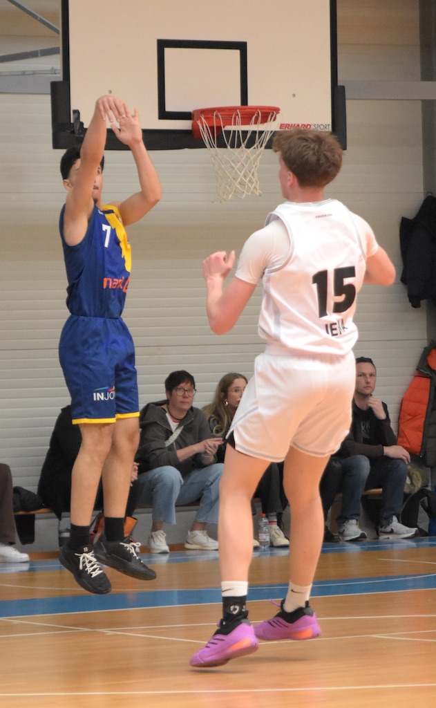 Ein Spieler in blau schießt einen Korb, während ein gegnerischer Spieler in weiß verteidigt.