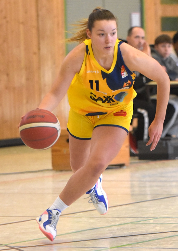 Basketballspielerin dribbelt konzentriert. Gelbes Trikot, blau-weiße Schuhe. Holzverkleidung im Hintergrund.