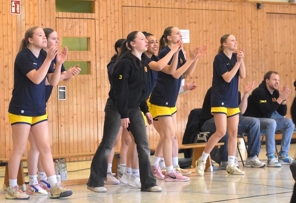 Eine Gruppe von jubelnden Frauen und einem Mann in blauen und gelben Sportkleidern auf einer Bank.