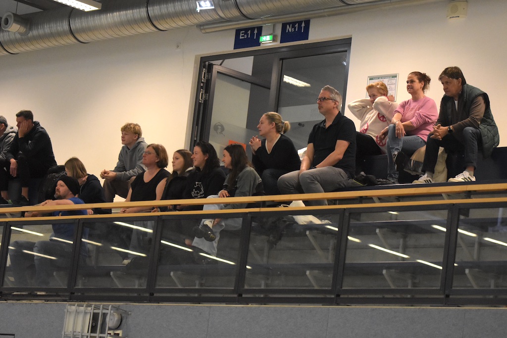 Zuschauergruppe auf Tribünen mit verschiedenen Emotionen, überwiegend in dunkler Kleidung.
