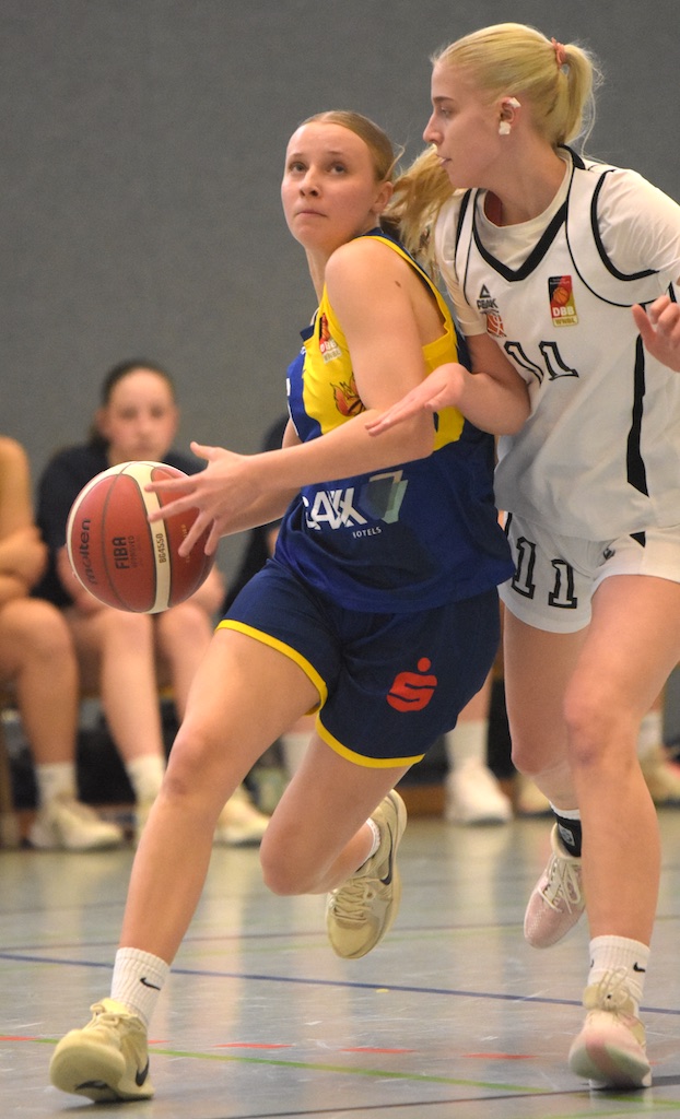 Eine Basketballspielerin in blau-gelbem Trikot versucht, an einer Gegenspielerin in Weiß vorbeizukommen.