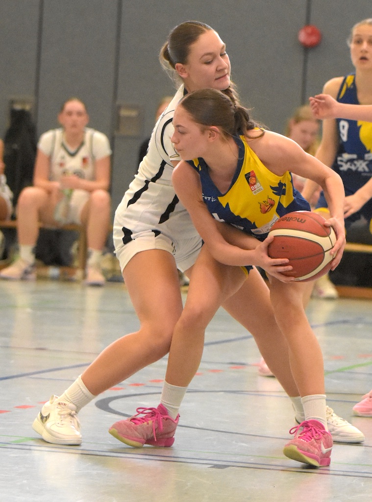 Zwei Basketballspielerinnen kämpfen um den Ball, eine im gelb-blauen Trikot, die andere in weiß.