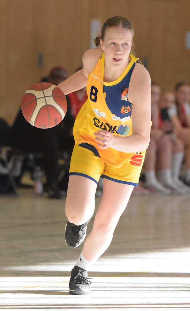 Eine Frau im gelben Basketballtrikot dribbelt konzentriert den Ball auf einem Hallenboden.