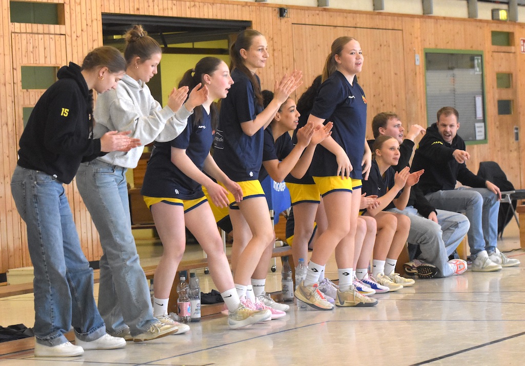 Frauen in Sportkleidung jubeln und klatschen auf einer Bank; hölzerne Sporthalle im Hintergrund.