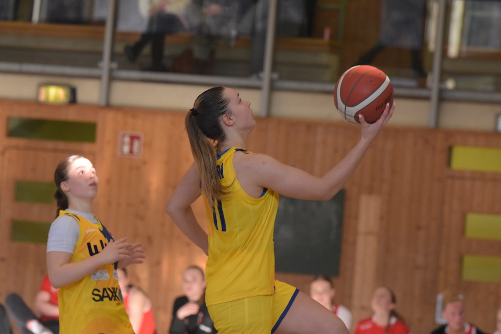 Basketballspielerin konzentriert beim Korbleger, gelbes Trikot, roter Ball, Holzwand im Hintergrund.