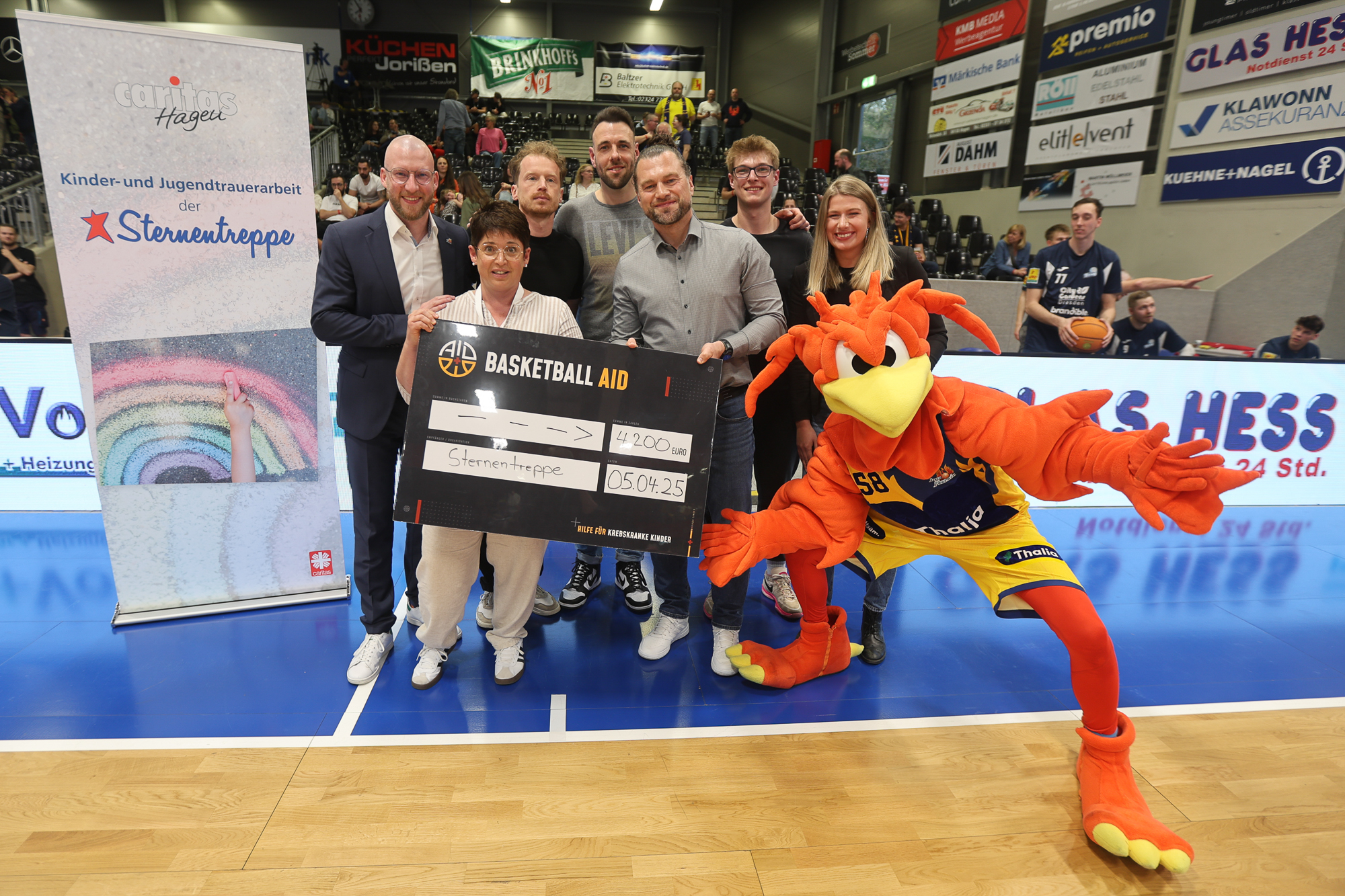 Eine Gruppe lächelt mit einem großen Scheck neben einem Basketball-Maskottchen in orangem Outfit.