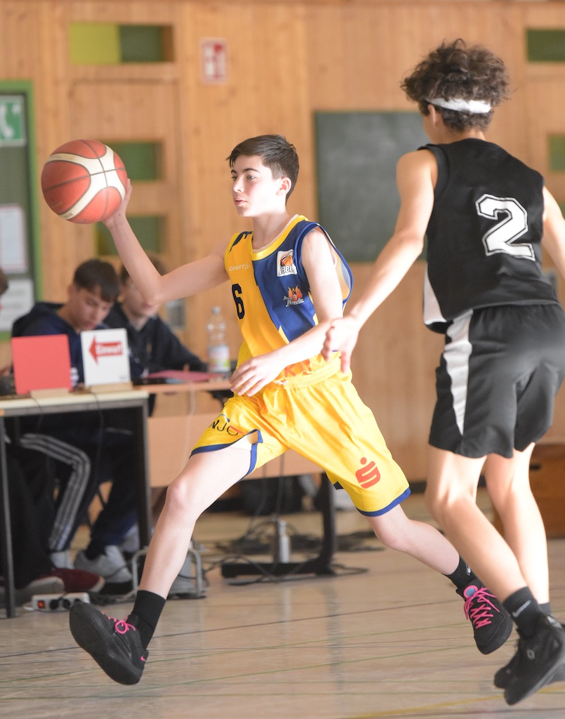 Ein Jugendlicher im gelben Trikot passt einen Basketball, während ein Gegner in Schwarz verteidigt.
