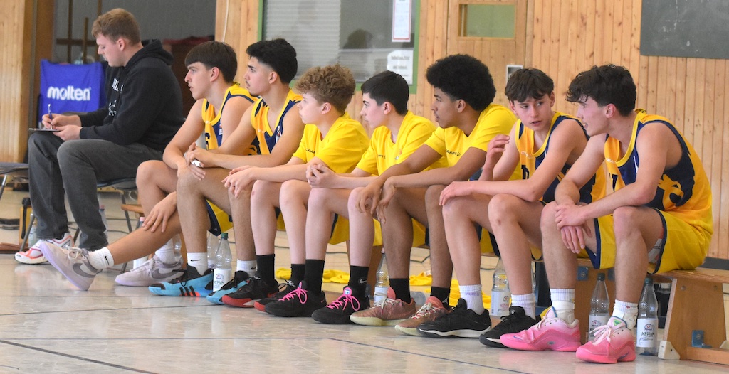 Eine Gruppe von acht jungen Männern in gelben Basketballtrikots auf einer Bank sitzt.