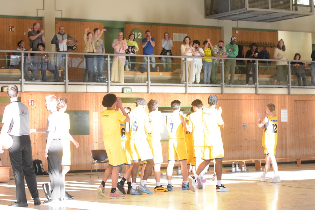 Eine Basketballmannschaft in gelben Trikots applaudiert; Zuschauer klatschen auf einer Tribüne.