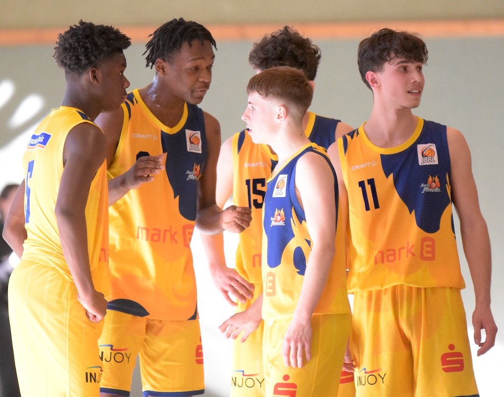 Fünf junge Basketballspieler in gelben Trikots, konzentriert, in einer Besprechung auf dem Spielfeld.