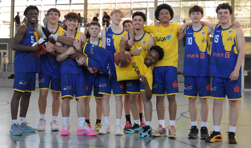 Eine Gruppe junger Basketballspieler in blauen Trikots, lachend und einen Spieler hochhebend.