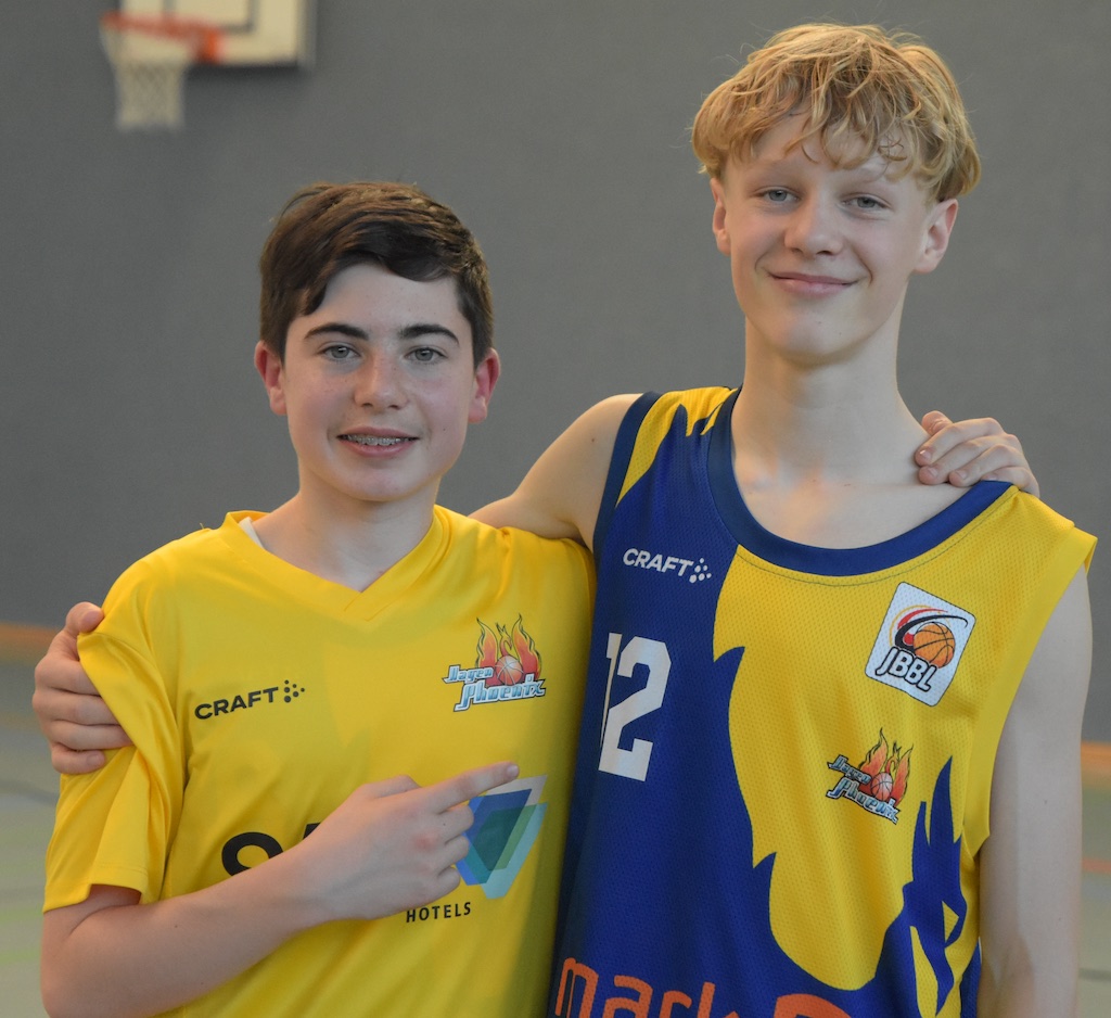 Zwei junge Basketballspieler lächeln; einer zeigt auf den anderen. In gelbem und blauem Trikot.
