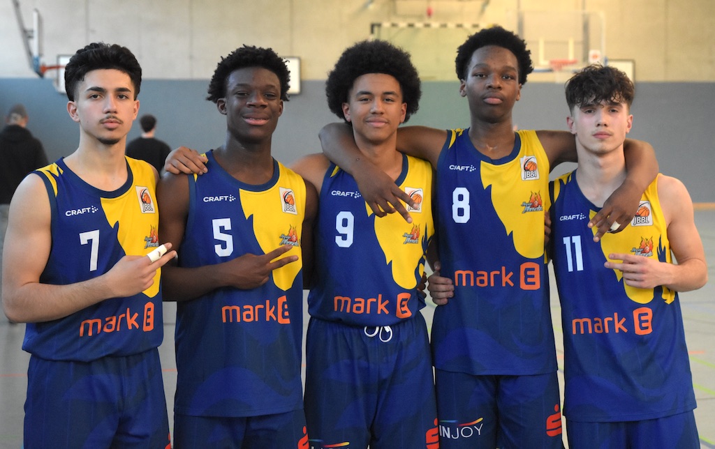 Fünf junge Basketballspieler in blau-gelben Trikots lächeln in die Kamera, Arme um die Schultern.