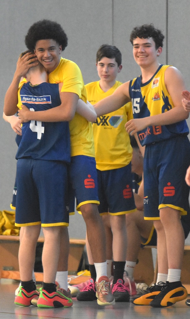 Vier junge Basketballspieler in gelben und blauen Trikots, lächeln und umarmen sich auf dem Spielfeld.
