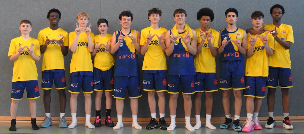 Elf Jugendliche im Basketball-Trikots lächeln und zeigen Handgesten vor grauer Wand.