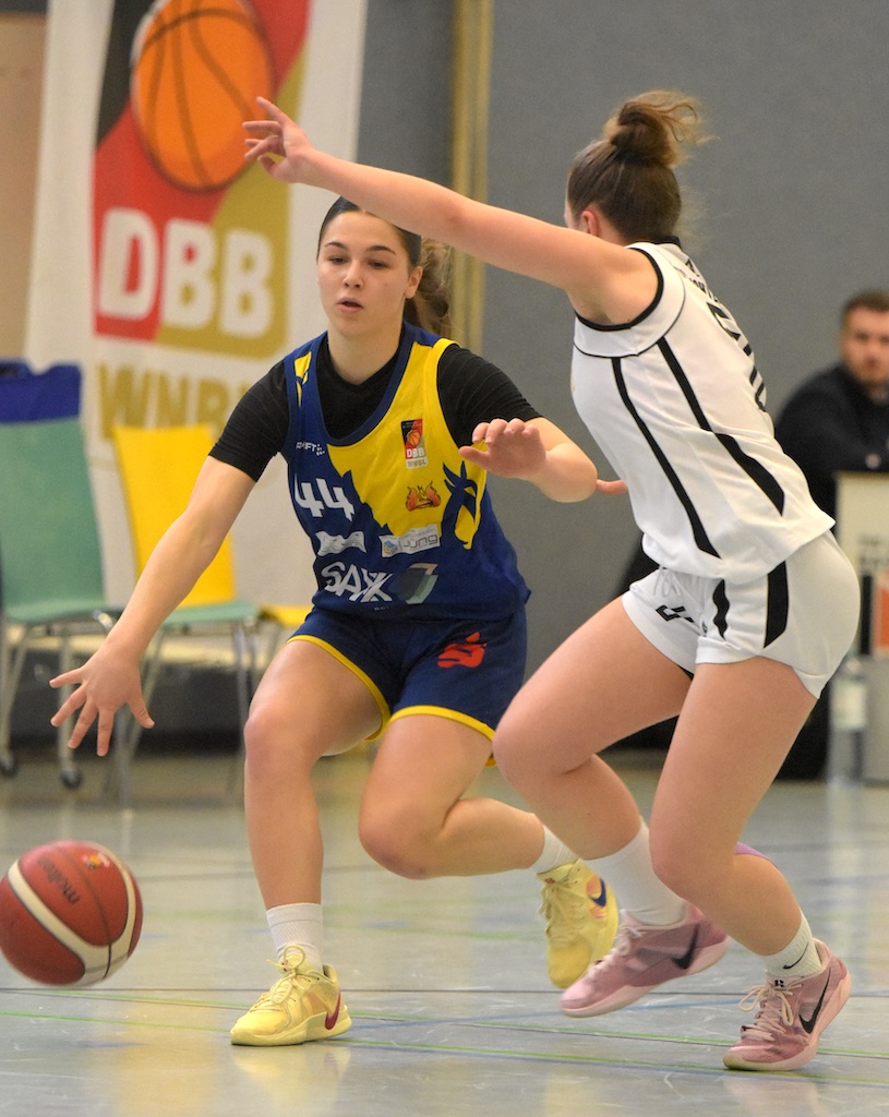 Zwei Basketballspielerinnen im Spiel. Eine dribbelt im gelben Trikot, die andere blockt im weißen.