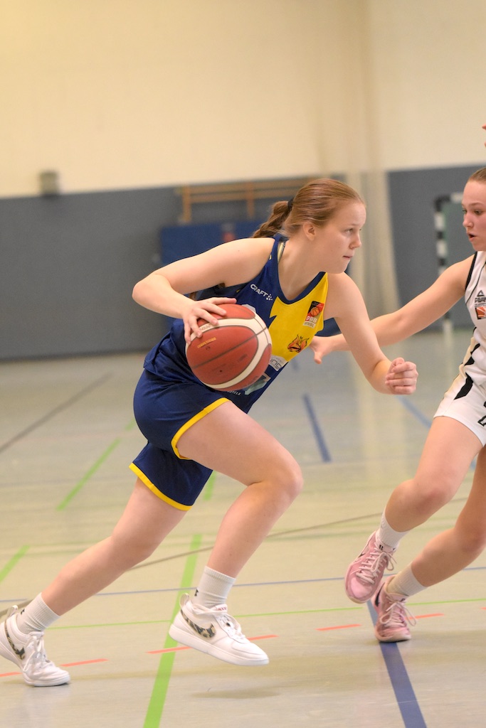 Basketballspielerin in blau-gelbem Trikot dribbelt fokussiert den Ball auf dem Hallenboden.