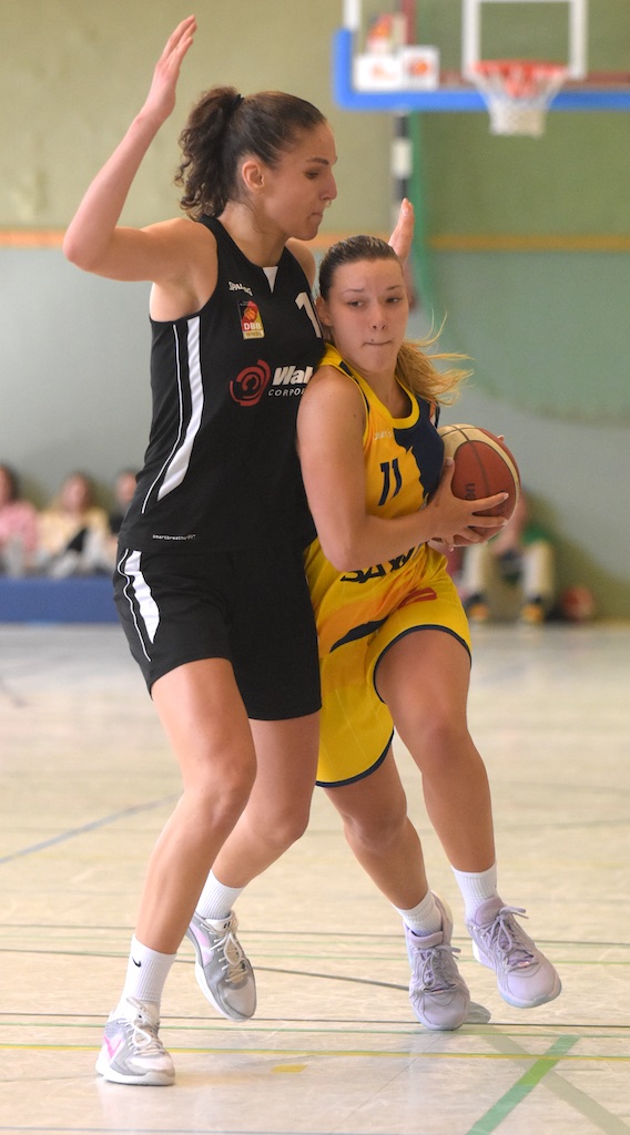 Basketballspielerin in gelbem Trikot dribbelt, wird von Spielerin in Schwarz verteidigt, konzentriert.