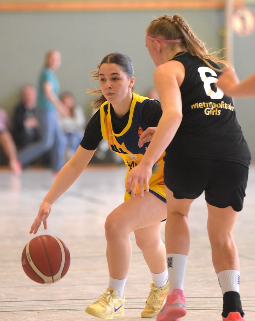 Eine Basketballspielerin in gelber Uniform dribbelt, verfolgt von einer Gegnerin in Schwarz.