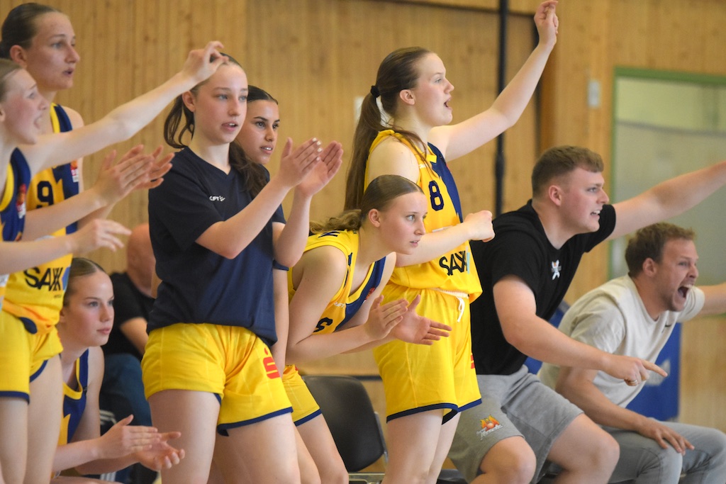 Basketball-Spieler:innen jubeln lebhaft, tragen gelbe Trikots und blaue Details, wirken begeistert.
