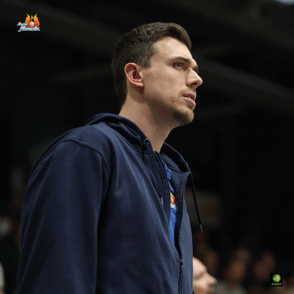 Mann mit ernstem Ausdruck in blauer Kapuzenjacke, dunkles Haar, konzentrierter Blick nach rechts.