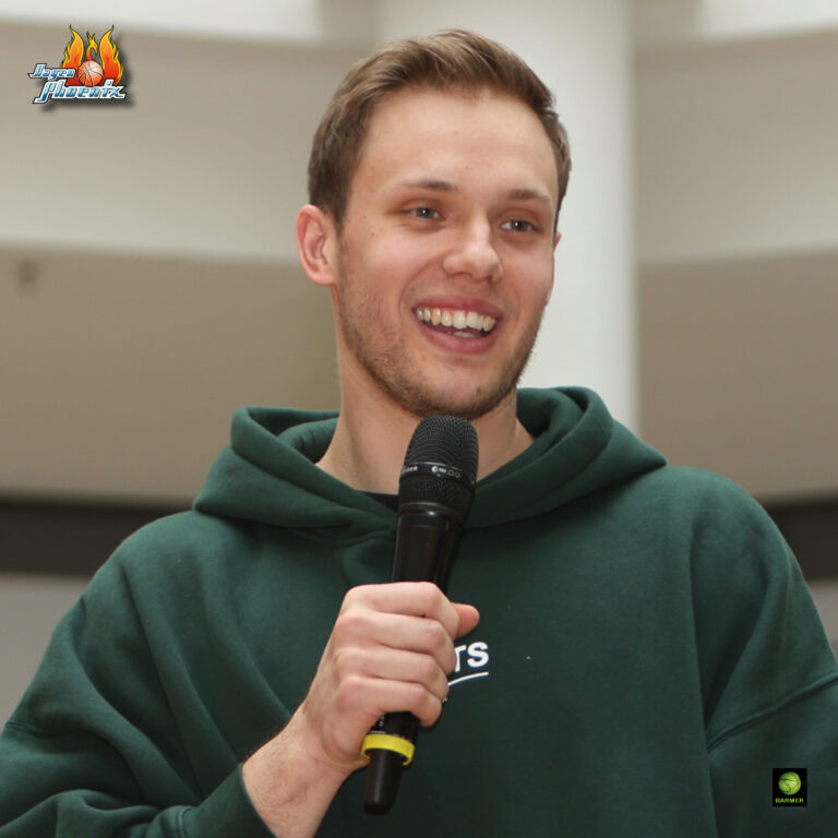 Ein lächelnder Mann hält ein Mikrofon und trägt einen dunkelgrünen Hoodie.