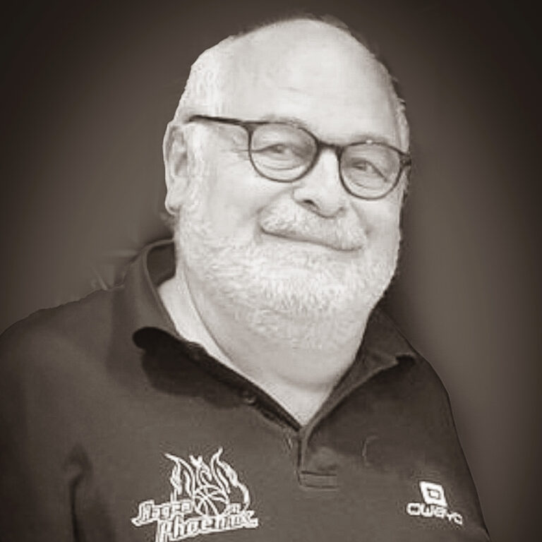 Älterer Mann mit Brille lächelt, trägt schwarzes Polo mit Logos, Hintergrund dunkel.