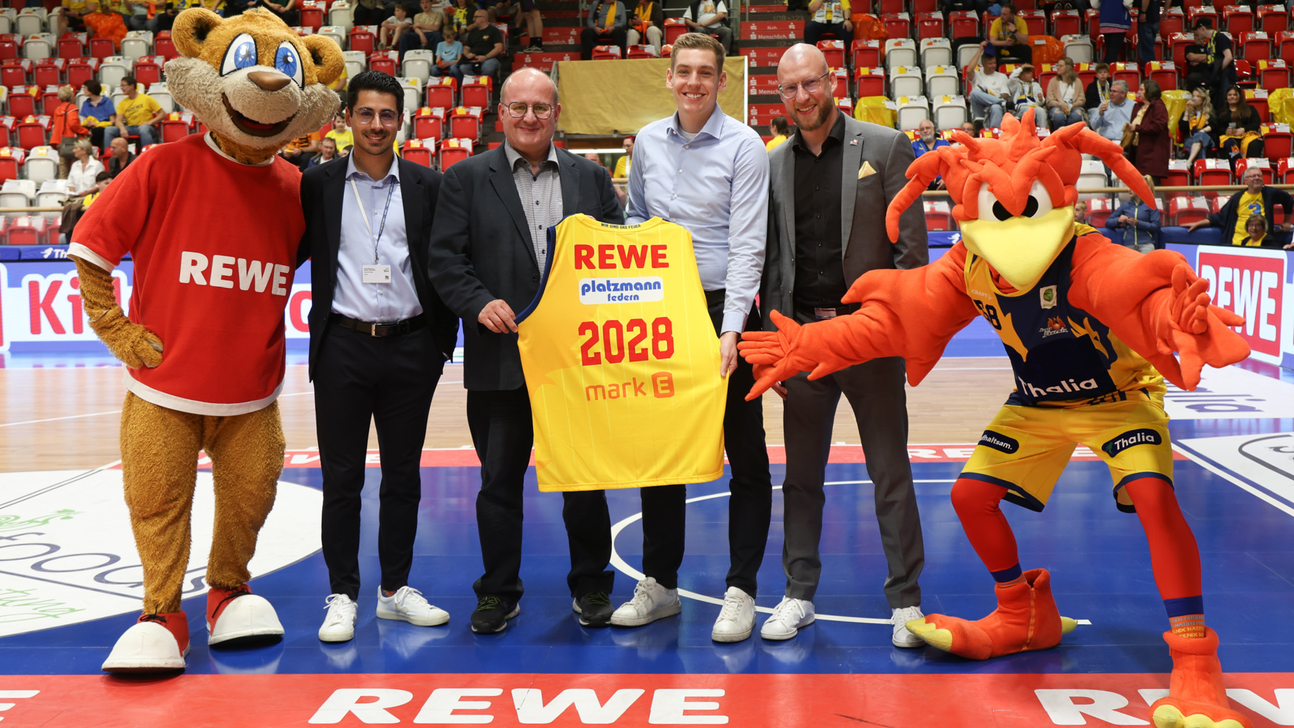 Eine Gruppe von sechs Personen, darunter zwei Maskottchen, hält ein gelbes Sporttrikot hoch.