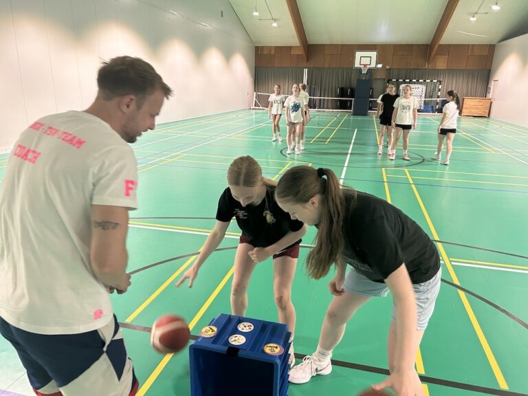 Eine Gruppe junger Leute spielt lachend in einer Turnhalle mit einem Ball.