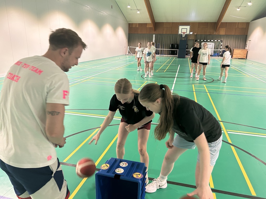 Eine Gruppe junger Leute spielt lachend in einer Turnhalle mit einem Ball.