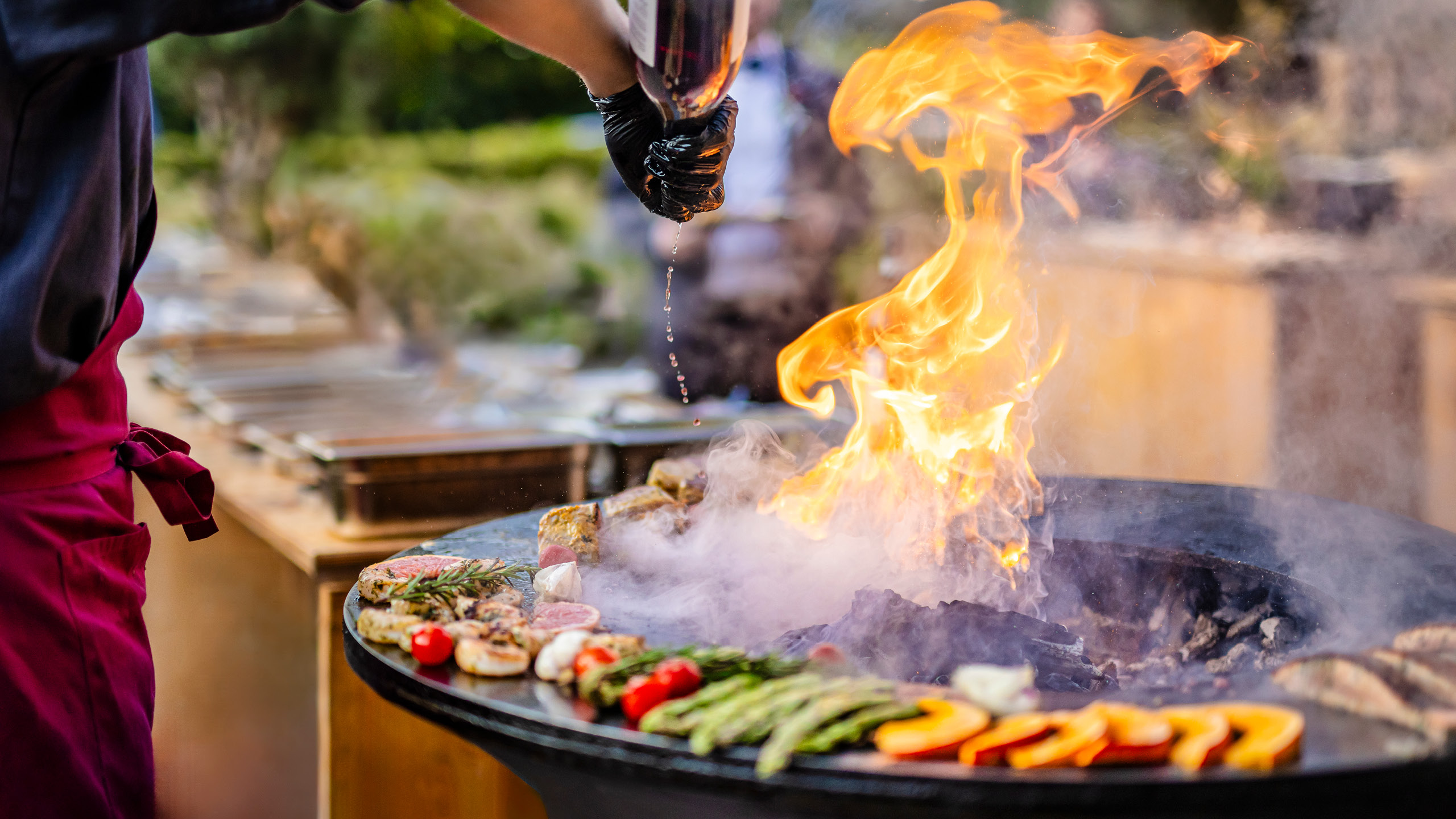 Eine Person grillt mit Flammen; bunte Gemüse im Vordergrund. Dynamische Szene mit intensivem Feuer.