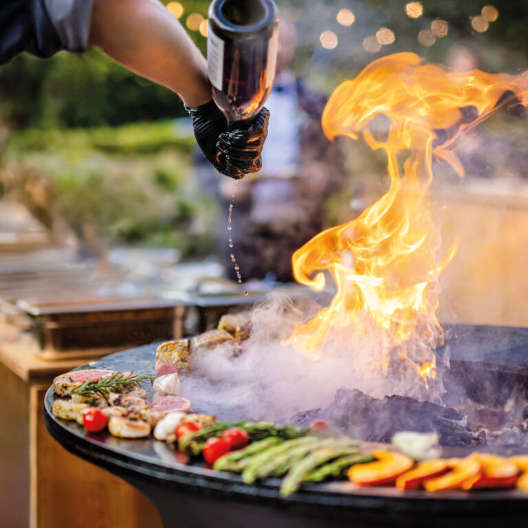 Eine Hand gießt Flüssigkeit auf Flammen über einem Grill mit Fleisch und Gemüse, funkelndes Bokeh.