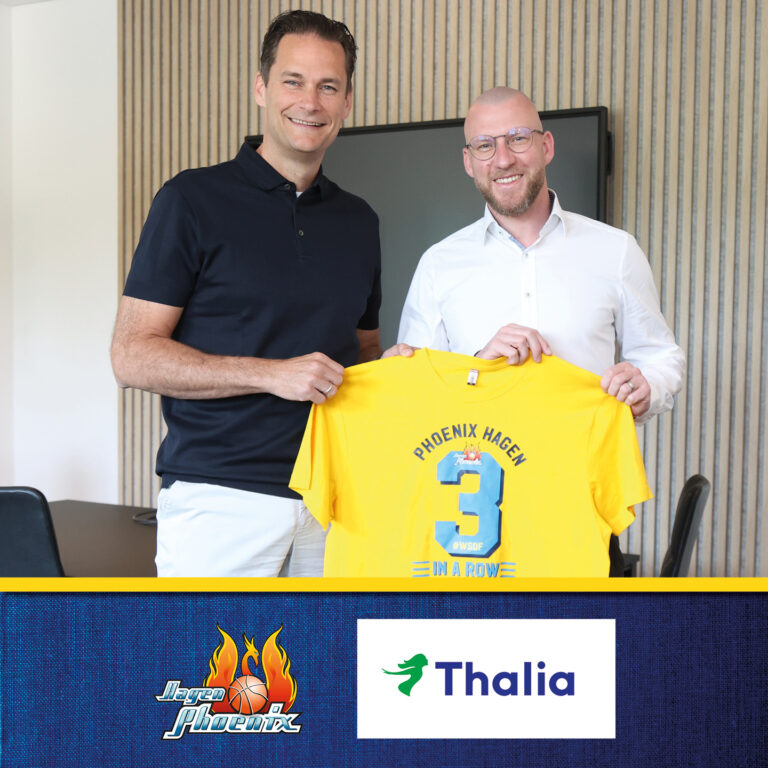 Zwei Männer lächeln, halten gelbes Phoenix Hagen T-Shirt, im Büro mit blauer Wand.