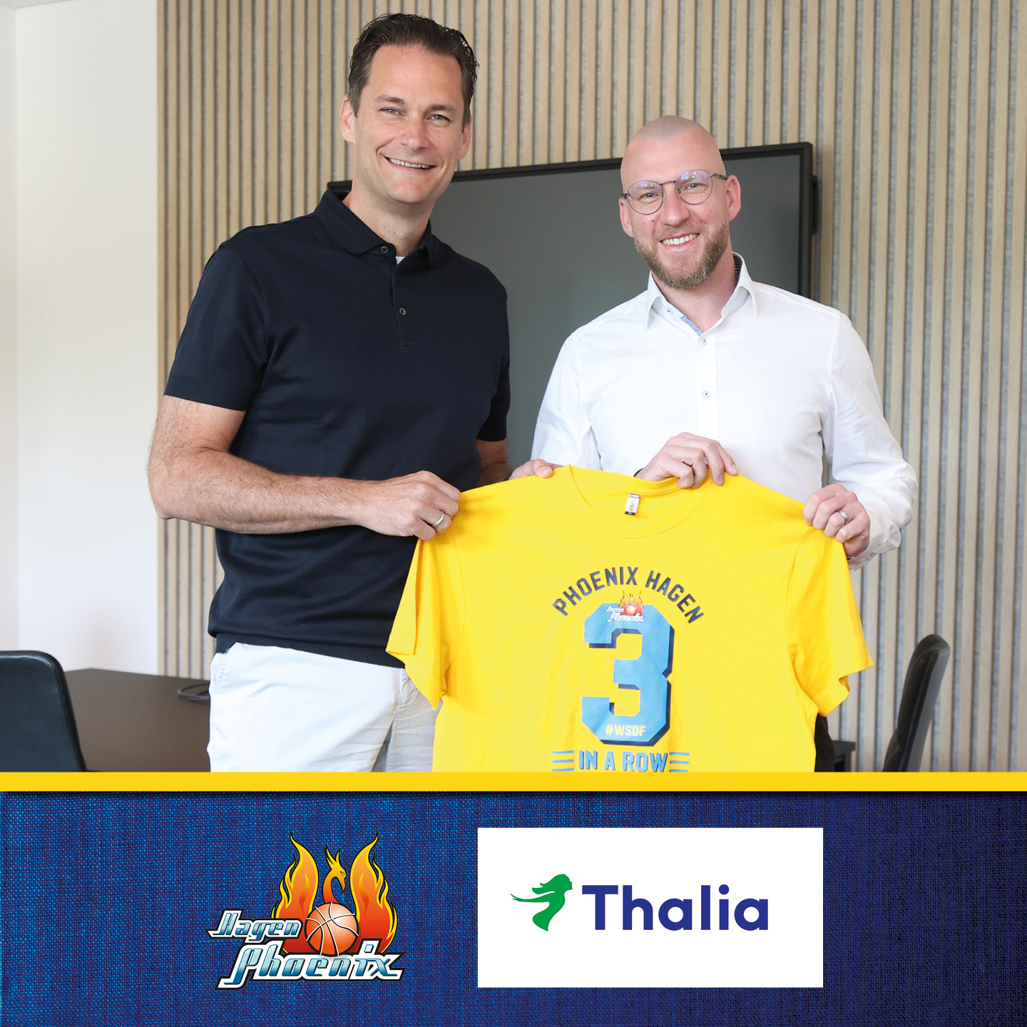 Zwei Männer lächeln, halten gelbes Phoenix Hagen T-Shirt, im Büro mit blauer Wand.