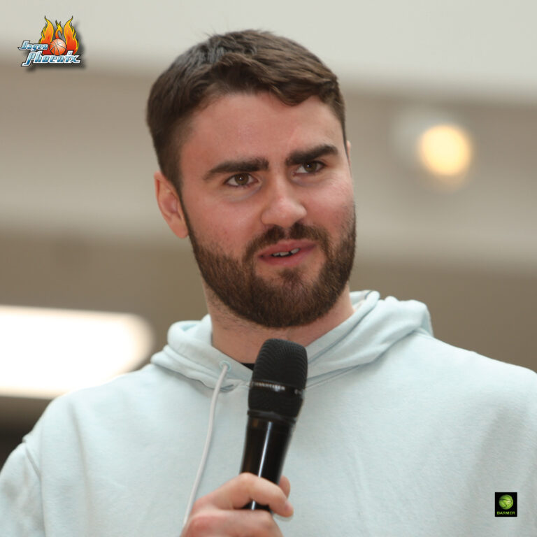 Ein Mann mit Bart in grauem Hoodie und Mikrofon, spricht konzentriert, Hintergrund unscharf.