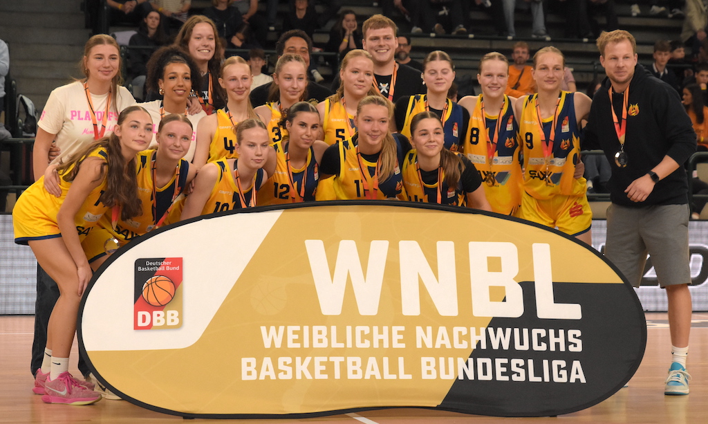 Eine Basketballmannschaft in gelben Trikots lächelt bei einer Siegerehrung in die Kamera.