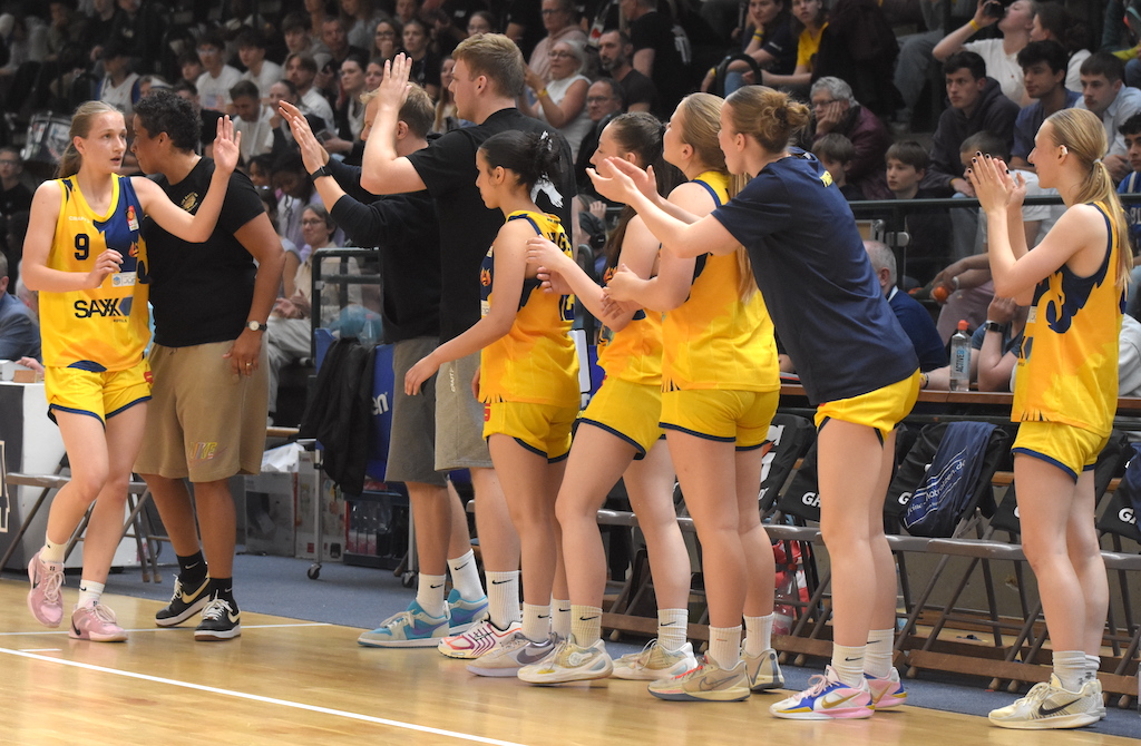 Eine Frau im gelben Basketballtrikot wechselt mit High-Five die Bank; Team jubelt aufgeregt.
