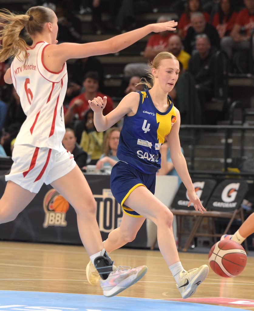 Basketballspielerin im blauen Trikot dribbelt konzentriert; Gegnerin im weißen Trikot verteidigt.