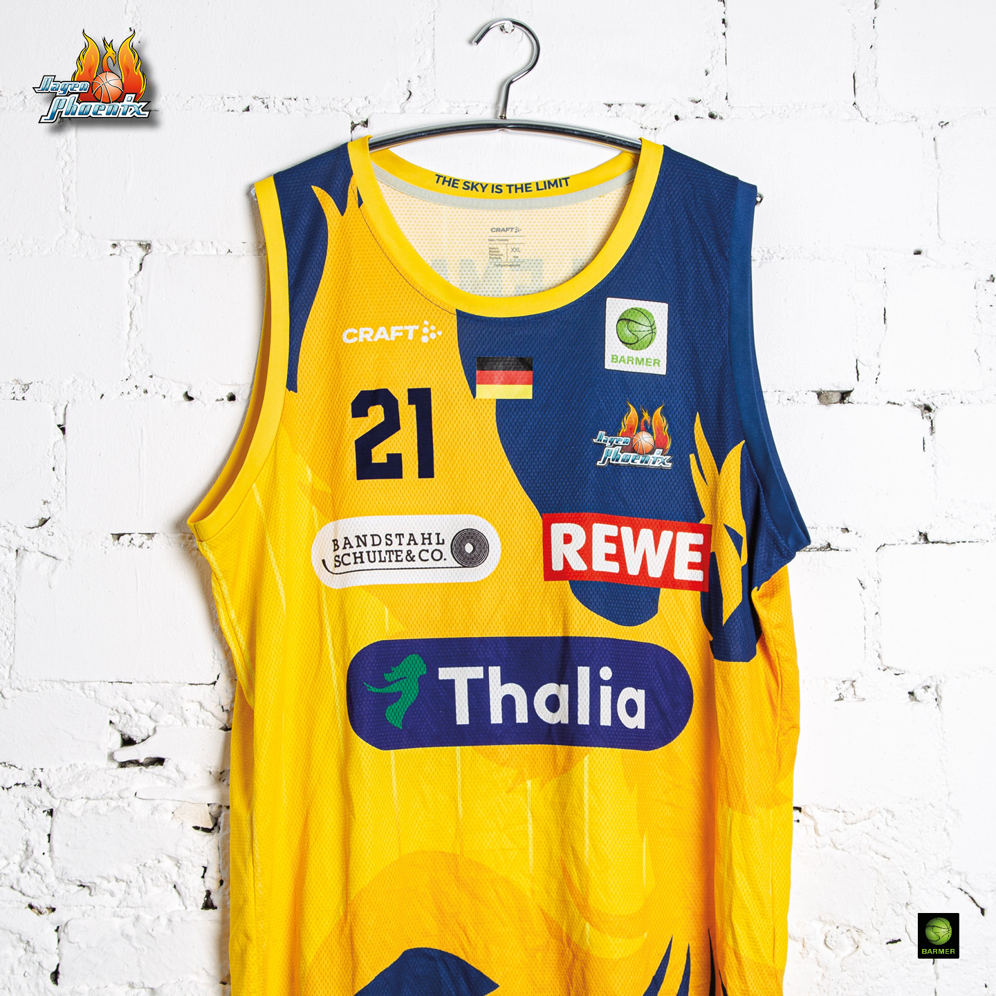 Ein gelbes und blaues Basketballtrikot mit "REWE" und "Thalia" Logos hängt an einer weißen Wand.